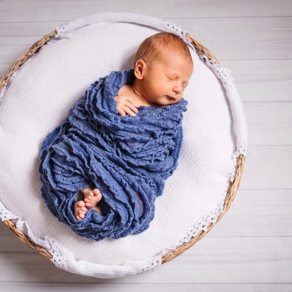 Newborn baby enveloped in blue scarf sleeps on white pillow